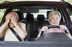A senior man covers his eyes in the passenger's seat as a senior woman drives.
