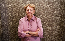 A senior women in front of a brown wall with white flower designs.