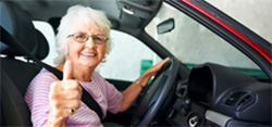 A senior woman behind the wheel gives a 'thumbs up'.